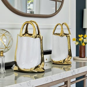 Peut inclure: Deux vases décoratifs en céramique blancs et dorés en forme de sacs à main. Les vases ont une finition brillante avec des poignées et des bordures dorées. Ils sont exposés sur une table en marbre, ajoutant une touche de luxe à la décoration.