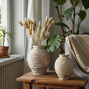 Può includere: Due vasi in ceramica beige strutturata con un design a coste su un tavolo di legno. Un vaso contiene erba coda di coniglio secca e soffice di colore marrone chiaro. Una piccola pianta in vaso e una pianta a foglie grandi sono sullo sfondo.