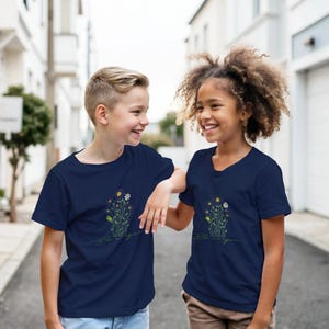 Könnte beinhalten: Zwei lächelnde Kinder tragen marineblaue T-Shirts mit einem Blumendesign. Das Design zeigt gelbe, weiße und grüne Blumen mit dem Text "Be Unique" in Schreibschrift. Sie stehen draußen.