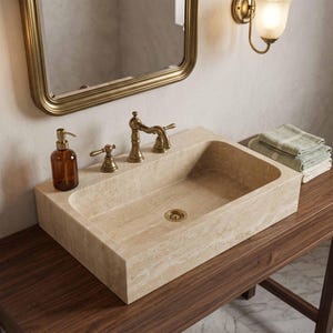 Puede incluir: Un lavabo rectangular de piedra beige con acabado texturizado natural, que cuenta con un grifo de latón con dos manijas a juego, un desagüe de latón y un dispensador de jabón de vidrio marrón, montado sobre un mueble de madera oscura.
