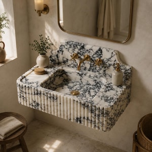 May include: A wall-mounted bathroom sink crafted from white marble with prominent black veining. The basin features a fluted exterior design, a gold-toned faucet with cross-handle controls, a matching gold drain, and a beige soap dispenser.