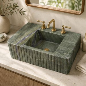 Può includere: Un lavabo rettangolare in marmo verde caratterizzato da un design esterno scanalato e una vasca interna liscia. Il lavabo è dotato di un rubinetto in oro spazzolato e due manopole laterali abbinate, montati sulla superficie in pietra.