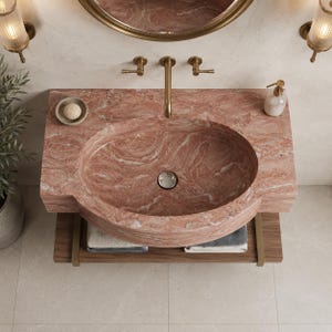 Puede incluir: Un mueble de ba&ntilde;o de m&aacute;rmol rosa con lavabo ovalado integrado, grifo de lat&oacute;n y dos mandos a juego. Debajo del lavabo hay un estante de madera con toallas dobladas. Un espejo redondo y dos apliques de pared est&aacute;n montados encima.
