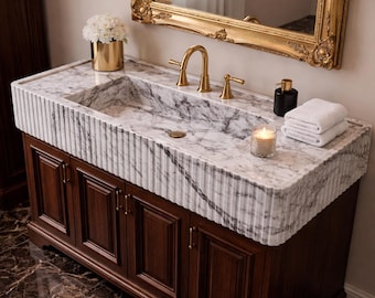 May include: A bathroom vanity featuring a white marble countertop with a fluted edge and an integrated sink. The base is made of dark brown wood with four cabinet doors and gold hardware. A gold faucet, a candle, and white towels are present.