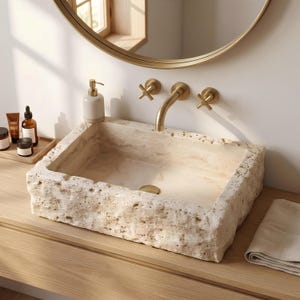 May include: A rectangular bathroom sink carved from beige travertine stone with a rough, natural exterior texture and a smooth interior basin. It features a wall-mounted brushed gold faucet with two cross-handle controls and a matching gold drain.