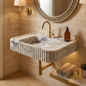 Op de afbeelding: Een aan de muur gemonteerde witte marmeren wastafel met een geribbeld exterieurontwerp, voorzien van een goudkleurige kraan en bijpassende leidingen. De wastafel bevindt zich onder een ronde spiegel met een gouden lijst en een wandlamp.