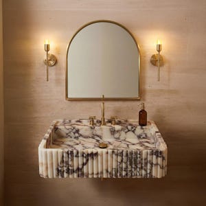 Puede incluir: Un lavabo rectangular de mármol montado en la pared con exterior estriado y vetas moradas, con grifo y desagüe dorados. Sobre el lavabo hay un espejo arqueado con marco dorado, flanqueado por dos apliques de pared dorados verticales.