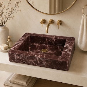 Puede incluir: Un lavabo rectangular fabricado en mármol color burdeos intenso con vetas blancas pronunciadas. La cubeta cuenta con una tapa de desagüe circular central en acabado latón cepillado, a juego con el grifo de pared y los mandos superiores.