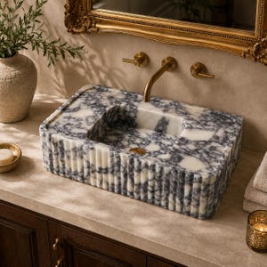 May include: A rectangular bathroom sink crafted from white and grey marbled stone, featuring a fluted exterior texture. The basin is equipped with a wall-mounted brass faucet and matching handles, set against a neutral wall with a gold-framed mirror.