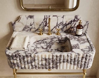 May include: A white and purple marble vanity sink featuring a fluted front design, a matching marble backsplash, and a brass faucet with two cross-handle knobs. A folded beige towel and a brown glass soap dispenser sit on the countertop.