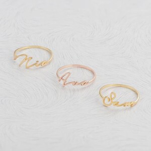 Può includere: Tre sottili anelli in metallo, ognuno con un nome diverso scritto in corsivo. Un anello è color oro, uno è color oro rosa e uno è color oro. I nomi visualizzati sugli anelli sono Nia, Ana e Sam in un carattere delicato.