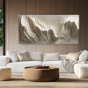 Op de afbeelding: Een groot, gestructureerd, abstract kunstwerk in tinten wit en crème hangt boven een witte bank. Het kunstwerk heeft een vloeiend, golfachtig ontwerp. De woonkamer omvat ook een ronde, bruine salontafel en een beige tapijt.