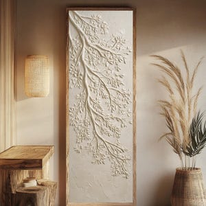 Puede incluir: Una obra de arte mural rectangular con un diseño de rama de árbol blanco en relieve. La obra de arte está enmarcada en un marco de madera de color claro. La habitación también cuenta con una lámpara tejida, taburetes de madera y una cesta tejida con plantas altas y secas.
