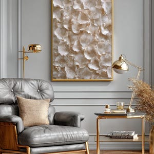 May include: A framed artwork with a gold border features a textured, abstract design in shades of beige and white. A gray leather armchair with a wooden frame and a gold side table with a black top are also visible. A gold lamp and a decorative pillow complete the scene.