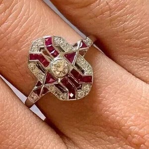 Peut inclure: Bague en argent au design géométrique. Elle présente un diamant rond central, entouré de pierres rectangulaires rouges et de petits diamants. La bague est portée au doigt.