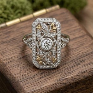 Puede incluir: Un anillo de plata adornado con un diamante redondo central, diamantes más pequeños y detalles dorados. El anillo presenta un diseño rectangular con motivos florales y de hojas. El anillo se muestra en un joyero de madera.