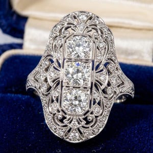 Op de afbeelding: Een sierlijke Art Deco-ring met drie centrale, rond geslepen diamanten in een gedetailleerde filigraan setting. De ring is gemaakt van zilverkleurig metaal en bevat talrijke kleinere diamanten. Gepresenteerd in een blauwe fluwelen ringdoos.