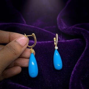 May include: Gold-coloured earrings featuring a teardrop-shaped, bright turquoise stone. The earrings have a small diamond accent and a hinged closure. The background is a deep purple velvet.