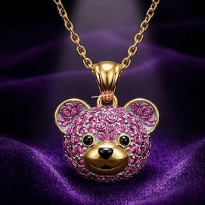 Könnte beinhalten: Eine goldfarbene Halskette mit einem Anhänger in Form eines Teddybären. Der Bärenkopf ist mit rosa Edelsteinen, schwarzen Augen und einer goldfarbenen Nase und Mund bedeckt. Rosenmuster verzieren die Ohren. Die Halskette hängt vor einem violetten Hintergrund.