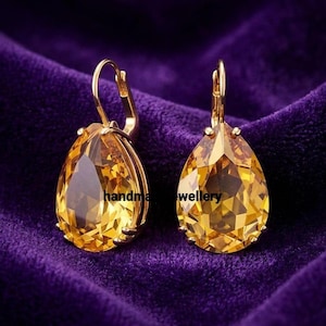 Peut inclure: Une paire de boucles d'oreilles dorées avec de grandes pierres précieuses jaunes en forme de goutte d'eau. Les boucles d'oreilles ont une fermeture à levier et sont posées sur un fond de velours violet foncé. Le texte "handma wellery" est visible.