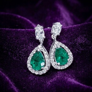 Peut inclure: Paire de boucles d'oreilles pendantes élégantes avec des émeraudes en forme de poire entourées d'un halo de petits diamants étincelants. Les boucles d'oreilles ont un accent de diamant en haut, sur fond de velours violet foncé.