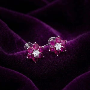 以下が含まれることがあります： 花型のスタッドピアス。各イヤリングは、6つの小さな鮮やかなピンク色の宝石に囲まれた中央の白い宝石を特徴としています。イヤリングはシルバーのポストにセットされ、濃い紫色のベルベットの表面に置かれています。