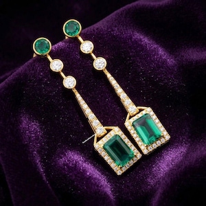 May include: A pair of gold earrings with emerald and diamond accents. Each earring features a large, rectangular emerald surrounded by a halo of diamonds, with a series of diamonds and emeralds ascending to the top. The earrings are set against a purple velvet background.