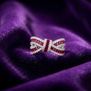 以下が含まれることがあります： 長方形の赤い宝石が中央に並び、小さな白い宝石が並んだリボン型のリング。深い紫色のベルベットの背景にセットされており、豪華でエレガントな美しさを演出しています。リングのデザインは複雑で人目を引きます。