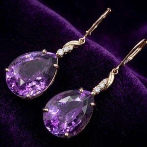 Può includere: Un paio di orecchini pendenti color oro con grandi pietre preziose viola a forma di pera. Ogni orecchino ha un piccolo gruppo di pietre preziose bianche e una chiusura a leva. Sfondo in velluto viola scuro.