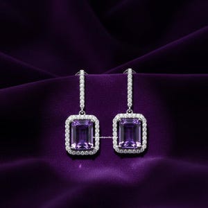 Può includere: Un paio di eleganti orecchini pendenti con pietre rettangolari viola scuro. Ogni orecchino è incorniciato da un alone di piccole pietre bianche scintillanti e sospeso da una delicata barra tempestata di diamanti. Su uno sfondo viola intenso.