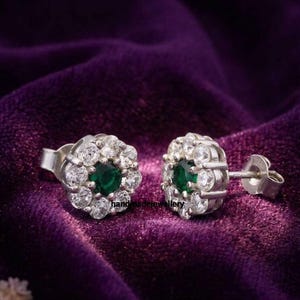 May include: Silver stud earrings with a floral design. Each earring has a central green gemstone encircled by smaller, clear gemstones. The earrings are set against a deep purple velvet background.