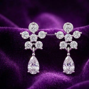 May include: A pair of silver-toned drop earrings with a floral design. Each earring features a cluster of round, sparkling stones and a pear-shaped, clear stone at the bottom. The earrings are set against a deep purple velvet background.