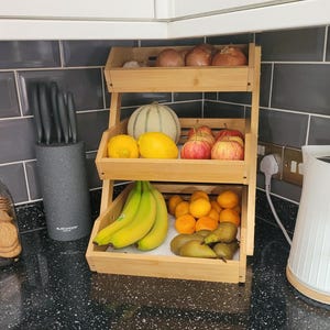 May include: A three-tiered bamboo fruit and vegetable stand filled with produce. The top shelf holds onions, the middle shelf has a melon, lemons, and apples, and the bottom shelf contains bananas, oranges, and pears.