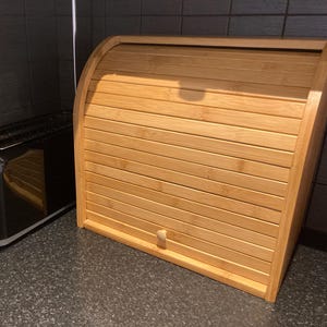 May include: A black two-slice toaster with chrome accents and a light-colored bamboo bread box. The bread box has a roll-top design and a wooden handle. The items are on a dark countertop.