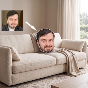 May include: A beige sofa with a large face-shaped pillow featuring a man's likeness. A smaller framed image of the same man is on the wall. A beige throw blanket is draped over the sofa. The room has a neutral color palette.