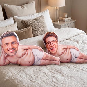Pode incluir: Duas almofadas de formato personalizado com rostos fotográficos impressos de duas pessoas diferentes em corpos vestindo roupa interior clara, posicionadas deitadas sobre uma cama com roupa de cama e almofadas em tons neutros.
