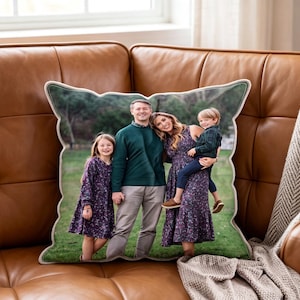 May include: A decorative pillow featuring a family portrait. The pillow has a white border and a green outline. The family of four is smiling and dressed in coordinated outfits. The pillow is on a brown leather couch.