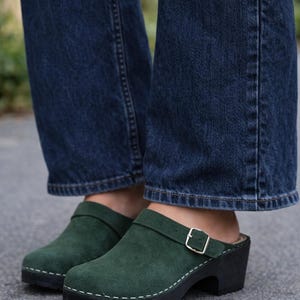 Op de afbeelding: Groene suède klompen met een gespriem en een zwarte zool. De klompen hebben een ronde neus en een kleine hak. De stiksels zijn wit. De klompen worden gedragen met een blauwe spijkerbroek.