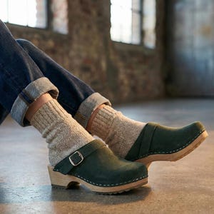May include: Dark green clogs with a buckle strap and wooden soles. The clogs are worn with beige knit socks and blue jeans. The shoes have a classic design with visible stitching around the sole.