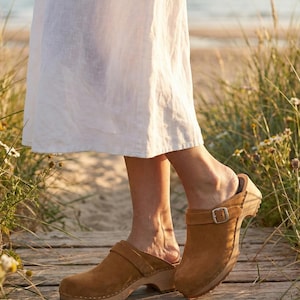 Può includere: Zoccoli in pelle scamosciata marrone con suole in legno e fibbie. Gli zoccoli sono indossati su una passerella di legno vicino alla spiaggia. L'immagine mostra gli zoccoli dalle caviglie in giù, con un vestito bianco.