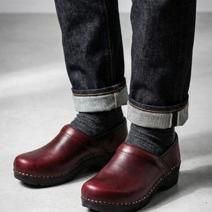 May include: A pair of burgundy leather clogs with a black sole, worn with gray socks and dark blue denim jeans. The jeans are cuffed at the ankles. The clogs have a classic, minimalist design.