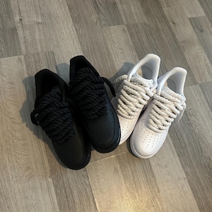 May include: Two pairs of sneakers, one black and one white, with thick, rope-like laces. The black sneakers are all black, while the white sneakers have white uppers and laces. The shoes are on a wooden floor.