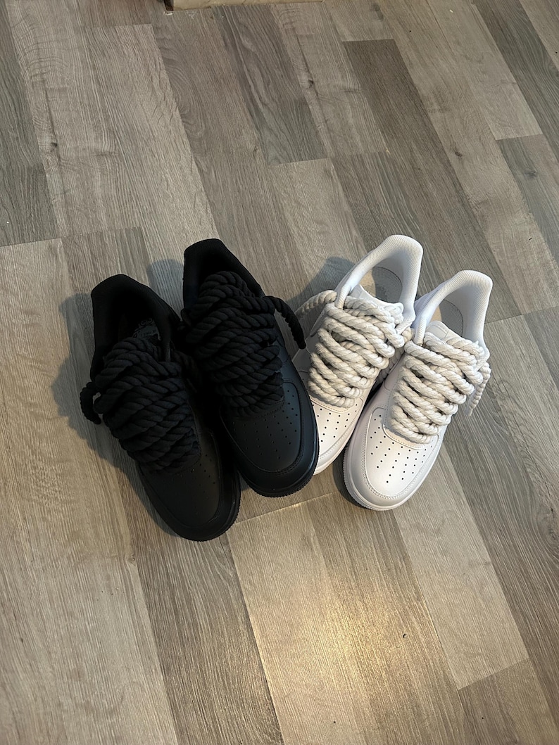May include: Two pairs of sneakers, one black and one white, with thick, rope-like laces. The black sneakers are all black, while the white sneakers have white uppers and laces. The shoes are on a wooden floor.