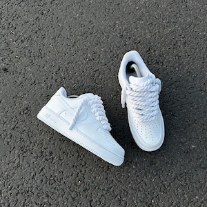 May include: A pair of white sneakers with thick, braided laces. The shoes have a classic design with perforations on the toe box and the word "AIR" on the side. The sneakers are set against a dark, textured surface.
