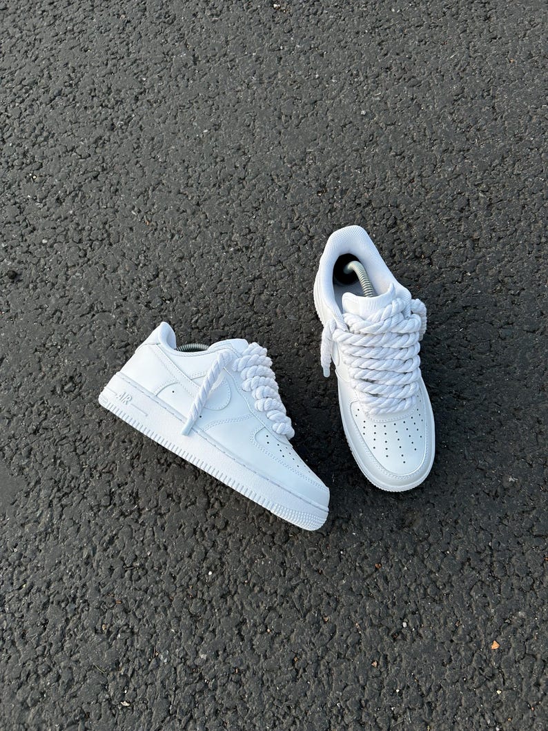 May include: A pair of white sneakers with thick, braided laces. The shoes have a classic design with perforations on the toe box and the word "AIR" on the side. The sneakers are set against a dark, textured surface.