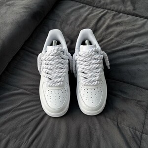 May include: A pair of white sneakers with thick, rope-like laces. The shoes are made of smooth leather and have a classic design with perforations on the toe box. They are set against a dark gray, textured background.