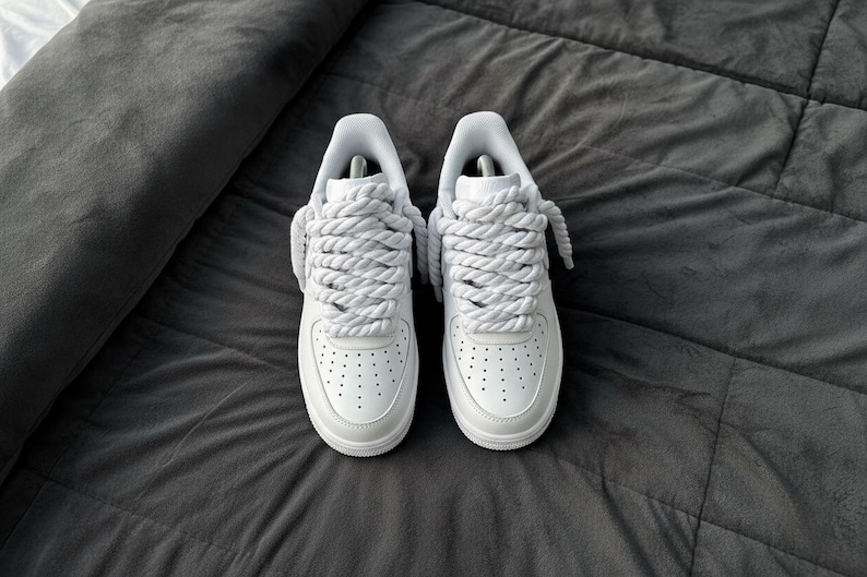 May include: A pair of white sneakers with thick, rope-like laces. The shoes are made of smooth leather and have a classic design with perforations on the toe box. They are set against a dark gray, textured background.