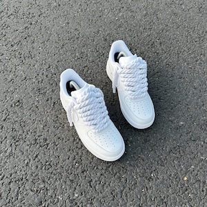 May include: A pair of white sneakers with thick, braided white laces. The shoes have a classic design with a perforated toe box and a white sole. The sneakers are set against a textured gray surface.