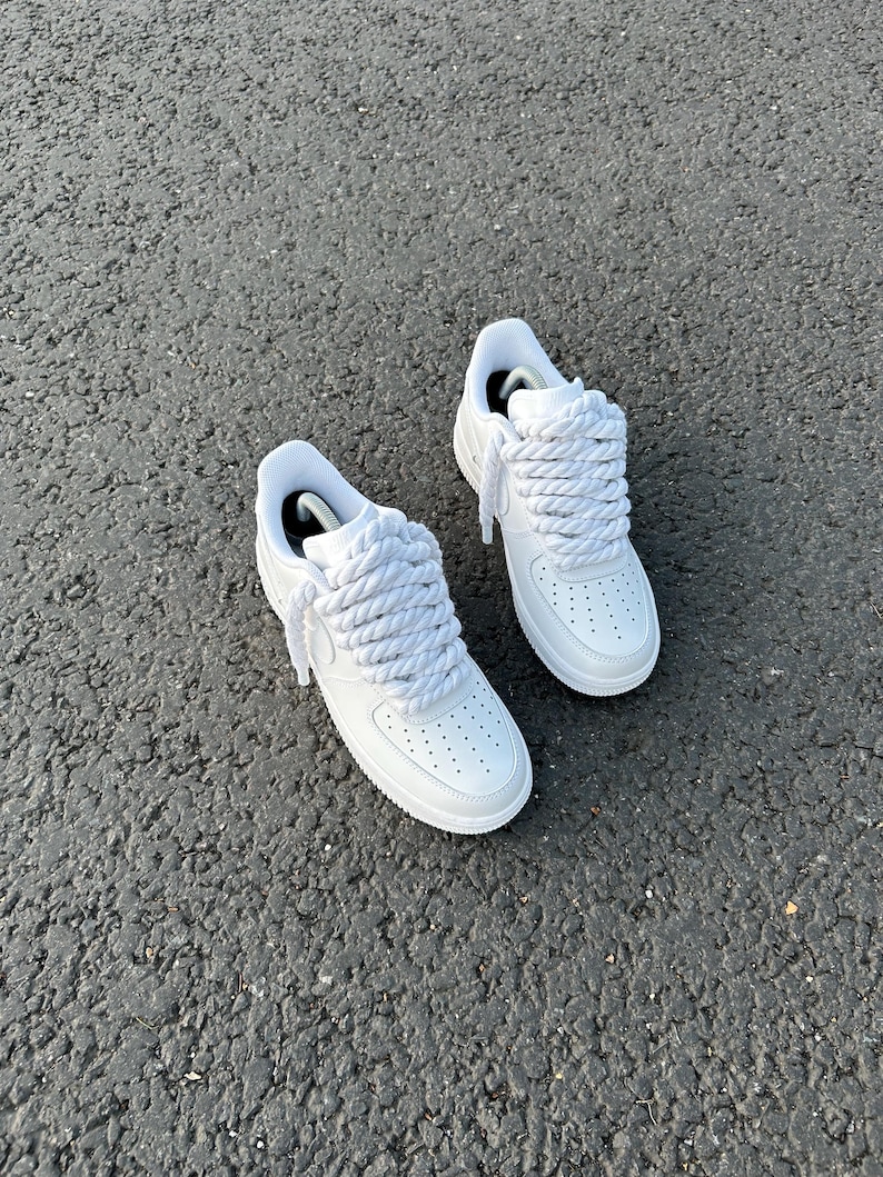 May include: A pair of white sneakers with thick, braided white laces. The shoes have a classic design with a perforated toe box and a white sole. The sneakers are set against a textured gray surface.