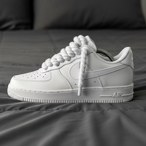 May include: A white leather sneaker with a classic design, featuring a perforated toe box, a prominent Swoosh logo, and thick white laces. The shoe is set against a gray background.